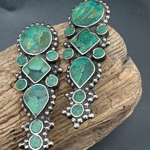 Pode incluir: Um par de brincos prateados com incrustações de pedras de cor turquesa. Os brincos apresentam uma série de formas geométricas, incluindo círculos, diamantes e gotas, rodeadas por uma borda de contas.