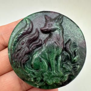 May include: A circular carved gemstone pendant featuring a fox sitting amidst stylized flames and floral elements. The stone displays a deep green base color with dark purple and black inclusions throughout the polished, textured surface.