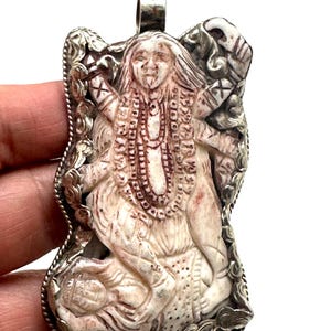 Puede incluir: Colgante tallado que representa una deidad con múltiples brazos, joyería intrincada y un rostro detallado. El colgante es de color crema con acentos marrón rojizo y un marco de metal plateado. Rectangular con un lazo en la parte superior.