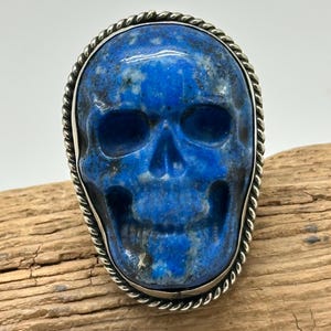 May include: A blue skull-shaped ring with a silver rope-like border. The skull is carved from a blue stone with white and gray marbling. The ring is set on a wooden surface.