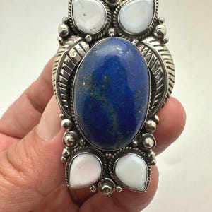 May include: A large statement ring featuring a central oval lapis lazuli stone surrounded by four teardrop-shaped white stones. The setting is crafted from polished silver with intricate leaf-like engravings, beaded accents, and twisted wire details.
