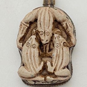 May include: A silver pendant featuring a detailed carving of three frogs, with one frog on top of the other two. The pendant has a gold-colored bail.