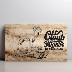 May include: Wooden wall art featuring a detailed illustration of a ram standing on a rock formation. The artwork includes the text "Climb Higher, Eat What Scares You" in a stylized font, set against a light brown wood grain background.