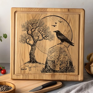 Op de afbeelding: Vierkante houten snijplank met een gegraveerd ontwerp van een boom, een raaf op een Keltisch kruis en een maan. De plank heeft een groef voor sappen langs de rand en is gemaakt van lichtgekleurd hout.