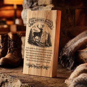 Op de afbeelding: Houten plaquette met de tekst "The Hunter's Prayer" en een hertenillustratie. De plaquette is gemaakt van lichtgekleurd hout en heeft gegraveerde tekst en een gedetailleerde afbeelding van een hert in een bosrijke omgeving. De plaquette staat rechtop.