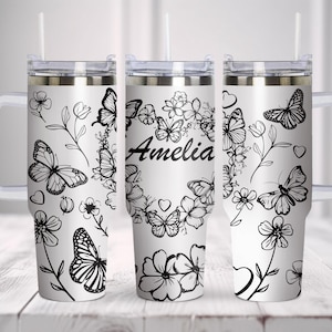 Puede incluir: Un vaso de acero inoxidable blanco con un diseño de mariposa y flores en blanco y negro. El vaso tiene una tapa y un asa. El texto "Amelia" está impreso en el vaso.