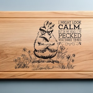 May include: Wooden cutting board with a carved design of an angry chicken and the text "I MIGHT LOOK CALM, BUT IN MY HEAD, I'VE PECKED YOU THREE TIMES." The board has a juice groove and is light brown.