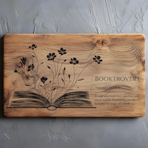 Può includere: Targa rettangolare in legno con la parola "BOOKTROVERT" e la sua definizione. Un motivo floreale nero emerge da un'illustrazione di libro aperto. La targa è realizzata in legno marrone chiaro con venature naturali.