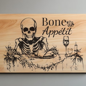 Puede incluir: Una tabla de cortar de madera con una ilustración en blanco y negro de un esqueleto sentado en una mesa con un plato de comida, una copa de vino y una vela. El texto "Bone Appétit" está escrito encima del esqueleto.