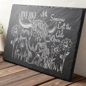 Puede incluir: Un letrero de pizarra negra con un diseño grabado en blanco de una vaca de las Tierras Altas con flores y el texto "Live like someone left the gate open."