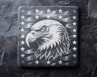 Puede incluir: Un posavasos de pizarra negra con una imagen grabada en blanco de la cabeza de un águila calva con el pico abierto, sobre un fondo de bandera estadounidense con estrellas.