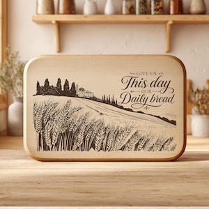 Op de afbeelding: Een rechthoekige snijplank van lichtgekleurd hout met een donkerbruine lasergravure van een korenveld, een gebouw in de verte en de tekst "GIVE US This day OUR Daily bread" met decoratieve krullen aan de rechterkant.