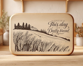 Puede incluir: Una tabla de cortar rectangular de madera clara con un grabado láser en marrón oscuro que muestra un campo de trigo, un edificio a lo lejos y el texto "GIVE US This day OUR Daily bread" con adornos decorativos a la derecha.
