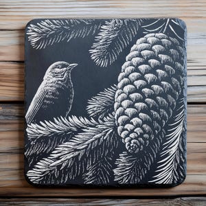 May include: Square, black slate coaster with a white, detailed illustration of a bird perched on a pine branch next to a pine cone. The coaster is set against a wooden background.