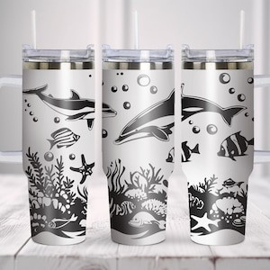 Puede incluir: Tres vasos de acero inoxidable con fondo blanco y siluetas negras de delfines, peces y corales. Los vasos tienen un asa y una tapa con una pajita.
