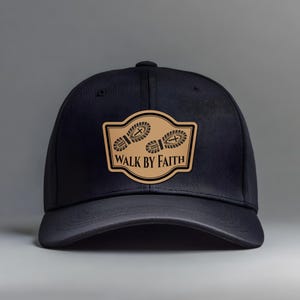May include: Black baseball cap with a curved brim. A tan patch on the front features two footprints and the words "WALK BY FAITH" in black lettering. The cap is made of a textured fabric.