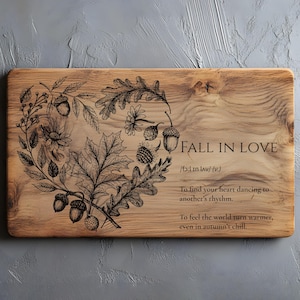 May include: A rectangular wooden chopping board with an engraved design. It features a wreath of leaves, acorns, and flowers, with the words "FALL IN LOVE" and a short poem. The wood has a natural grain and is light brown.