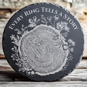 May include: A round, black slate coaster with the engraved text "EVERY RING TELLS A STORY". The coaster features a detailed tree ring design in the center, surrounded by floral and botanical illustrations. The coaster is a decorative item.