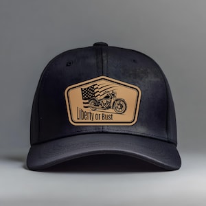 Può includere: Cappellino da baseball nero con toppa in pelle marrone. La toppa raffigura una motocicletta, la bandiera americana e la scritta "Liberty or Bust". Il cappellino ha una visiera curva e una corona strutturata.