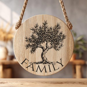 May include: A circular light-colored wooden plaque featuring a black ink illustration of an olive tree with a twisted trunk. The word FAMILY is printed in black capital letters below the tree. The plaque hangs from a thick, braided jute rope.