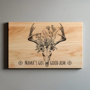 May include: A rectangular wooden sign with a deer skull design. The skull is adorned with flowers and antlers. The text "MAMA'S GOT GOOD AIM" is printed below the skull in black. The sign has a light wood grain.