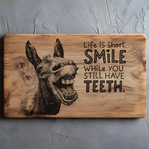 以下が含まれることがあります： ユーモラスなロバのイラストと「Life is Short. SMILE WHILE YOU STILL HAVE TEETH.」のテキストが描かれた長方形の木製まな板。ロバは大きく笑っています。木目が確認できます。