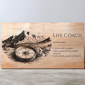 以下が含まれることがあります： 「LIFE COACH」という言葉と定義が書かれた木製の看板。コンパスと山脈の詳細なイラストが特徴です。テキストは「鏡とコンパス。行き詰まりから抜け出し、解放し、成長するのに役立ちます。」と書かれています。