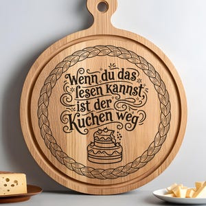 Peut inclure: Planche de service ronde en bois avec une poignée et une bordure décorative. La planche présente un texte gravé en allemand : "Wenn du das lesen kannst ist der Kuchen weg" et une illustration de gâteau.