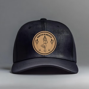 Puede incluir: Una gorra de béisbol negra con un parche circular de color tostado en la parte delantera. El parche muestra una ilustración de líneas negras de una persona en postura de yoga del árbol, rodeada de motivos florales y el texto "ROOTED IN BREATH".