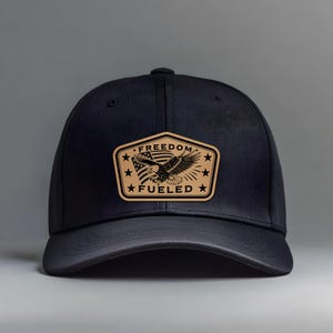 Może przedstawiać: Czarna czapka bejsbolowa ze skórzaną naszywką z orłem, amerykańską flagą i napisem "FREEDOM FUELED". Naszywka jest w kolorze brązowym z czarnym tekstem i wzorem. Czapka ma klasyczny sześciopanelowy design.