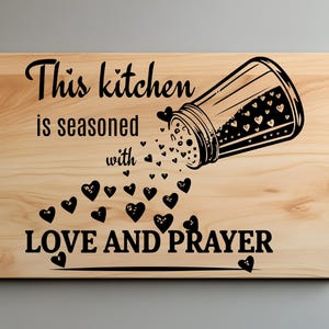 Puede incluir: Una tabla de cortar de madera con el texto "This kitchen is seasoned with LOVE AND PRAYER" y una ilustración en blanco y negro de un salero que vierte corazones.
