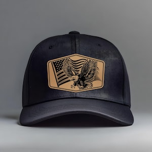 May include: A black baseball cap featuring a tan leather patch with an American flag and a detailed eagle illustration. The cap has a curved brim and a structured crown, suitable for outdoor wear.