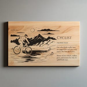 Può includere: Targa in legno con illustrazione di ciclista e testo. L'immagine in bianco e nero raffigura un ciclista in mountain bike davanti a una catena montuosa. Il testo include la definizione di "ciclista" e una citazione sul ciclismo.
