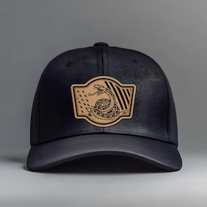 May include: Black baseball cap with a tan patch depicting a coiled snake, stars, and stripes. The patch is rectangular with a curved top and bottom. The cap has a curved peak and a button on top.