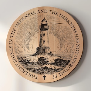 May include: Round wooden wall art featuring a detailed engraving of a lighthouse on a rocky island, surrounded by waves and radiating light. The text "THE LIGHT SHINES IN THE DARKNESS, AND THE DARKNESS HAS NOT OVERCOME IT" is etched around the edge, with a cross at the bottom.