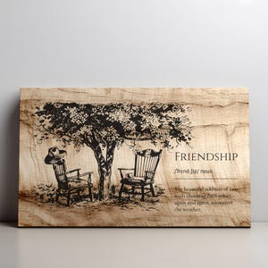 Puede incluir: Arte de pared de madera con una ilustración negra de dos sillas bajo un árbol. Un sombrero descansa sobre una silla. La palabra "FRIENDSHIP" y una definición están impresas en la madera.