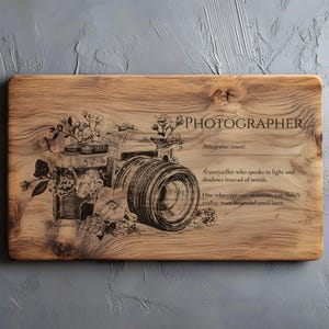 May include: Wooden cutting board with a detailed engraving of a vintage camera, adorned with floral accents. The word "PHOTOGRAPHER" is etched above the camera, along with a definition. The board has a natural wood grain finish.
