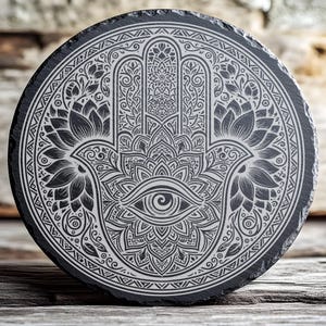 May include: Round black slate coaster with a detailed white engraved Hamsa hand design. The design includes an eye, lotus flowers, and intricate patterns. The coaster is placed on a wooden surface.