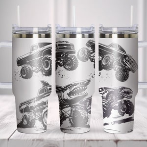 May include: Three stainless steel travel mugs with a white background and black graphic designs of monster trucks and sharks. The mugs have a handle and a lid with a straw.