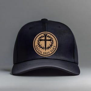 May include: A black baseball cap with a circular emblem. The emblem features a cross, nails, and a crown of thorns, with the words "SAVED, NOT SOFT" encircling the design. The cap is made of a dark material.