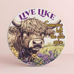 May include: A round white sign with a brown and white Highland cow in a field of purple and yellow flowers. The sign says "Live Like Someone Left The Gate Open" in purple and gold lettering.