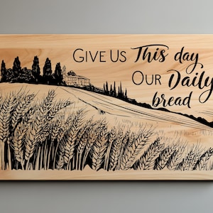 May include: A wooden sign with a black and white illustration of a field of wheat and a farmhouse in the distance. The text "GIVE US THIS DAY OUR DAILY BREAD" is written in a cursive font.