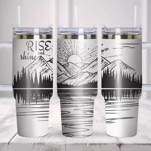May include: White travel tumblers with black and white mountain and lake designs. One tumbler has the text "Rise and Shine". Each tumbler has a handle, a clear lid, and a straw.
