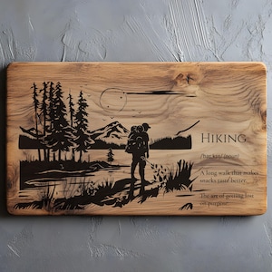 Puede incluir: Tabla de cortar rectangular de madera con un diseño de senderismo grabado. El diseño presenta un excursionista, árboles, montañas y la palabra "HIKING" con una definición y una cita. La tabla tiene un veteado natural.