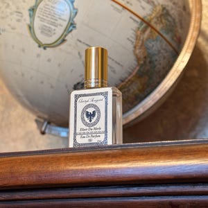 Op de afbeelding: Een heldere glazen parfumfles met een gouden dop en een etiket met de tekst "Elixir Du Merle Eau De Parfum". De fles staat voor een vintage wereldbol.