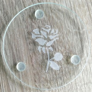 Könnte beinhalten: Ein klarer Glasuntersetzer mit einer geätzten Rose in der Mitte. Der Untersetzer hat drei kleine, klare, abgerundete Füße. Das Rosendesign zeigt detaillierte Blütenblätter und Blätter. Der Untersetzer ist rund.