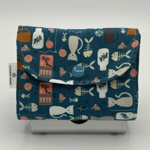 May include: A teal fabric wallet with a flap closure, adorned with a repeating pattern of white cat silhouettes, fish bones, milk bottles, and yarn balls. The wallet has a blue button closure and a small tag that says "MOMMA MAIN".