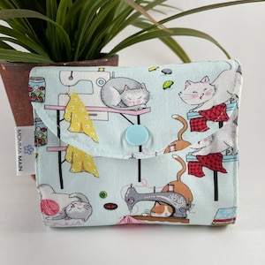 May include: A light blue fabric pouch with a snap closure. The pouch features a repeating pattern of cats sewing, with a sewing machine, yarn, and buttons. The text "MOMMA MAIN" is printed on the side of the pouch.