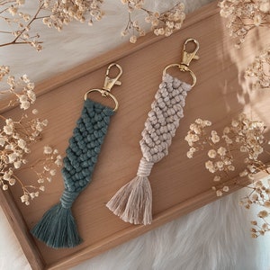Könnte beinhalten: Zwei Makramee-Schlüsselanhänger mit goldfarbenen Verschlüssen. Einer ist teal, der andere beige. Jeder hat ein geflochtenes Design und eine Quaste am unteren Ende. Sie liegen auf einem Holztablett mit Trockenblumen.
