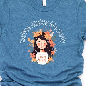 Puede incluir: Camiseta azul con mangas enrolladas. El gráfico muestra a una mujer sonriente con una corona de flores, sosteniendo una taza de café. El texto "Coffee Makes Me Smile" está encima de la ilustración. La taza de café tiene el texto "Siompe Nimpe".
